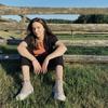 A woman sits on grass in front of a wooden fence and blue sky.